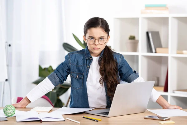 Sérieux enfant dans lunettes regarder caméra près ordinateur portable dans le bureau — Photo de stock