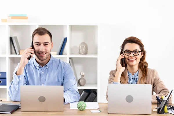 Exitoso hombre de negocios y mujer de negocios hablando en teléfonos inteligentes cerca de computadoras portátiles en la oficina - foto de stock