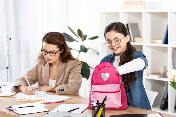 Fröhliches Kind blickt mit Brille im Büro auf Rucksack neben Mutter — Stockfoto