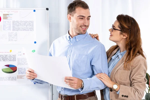 Cheerful woman in glasses looking at handsome man with blank paper near white board with charts and graphs — Stock Photo