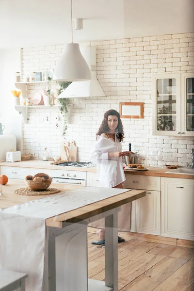 Mujer bonita en camisa blanca mirando hacia otro lado mientras se prepara el desayuno en la cocina - foto de stock