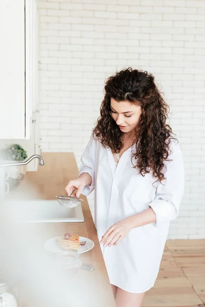 Радостная молодая женщина в белой рубашке готовит блины на кухне — стоковое фото