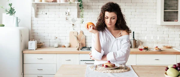 Panoramic shot of girl holding orange and looking at pancakes in kitchen — Stock Photo