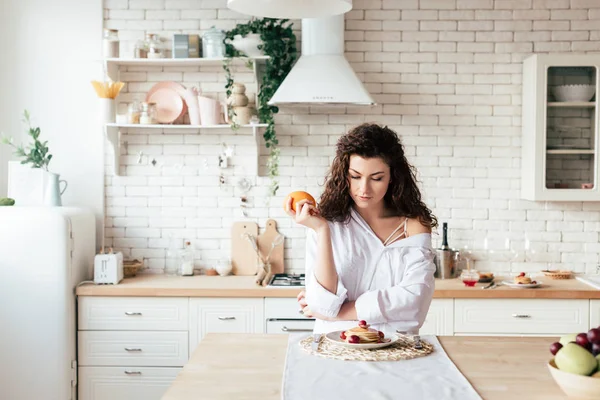 Plano panorámico de niña sosteniendo naranja y mirando panqueques en la cocina - foto de stock