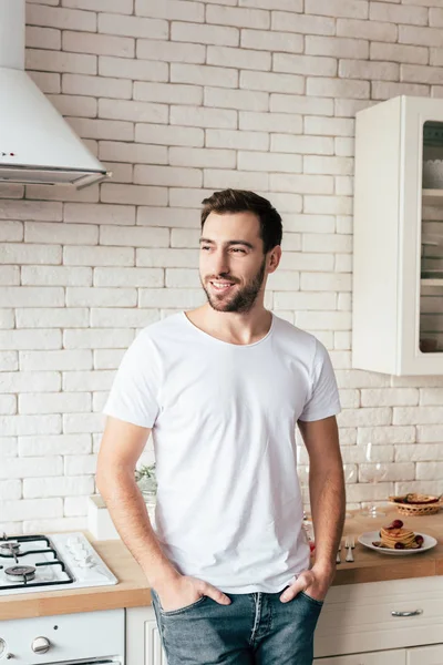 Uomo sorridente in jeans in piedi con le mani in tasca e distogliendo lo sguardo in cucina — Foto stock