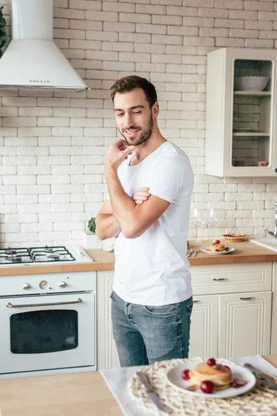 Homem barbudo em jeans olhando para panquecas com sorriso na cozinha — Fotografia de Stock