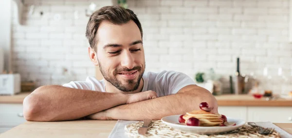 Panoramaaufnahme eines lächelnden bärtigen Mannes, der Pfannkuchen in der Küche betrachtet — Stockfoto