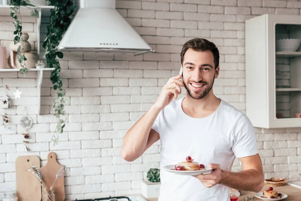 Lächelnder Mann spricht auf Smartphone und hält Teller mit Pfannkuchen in der Hand — Stockfoto