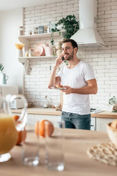 Hombre barbudo hablando en smartphone con sonrisa y sosteniendo plato con panqueques - foto de stock