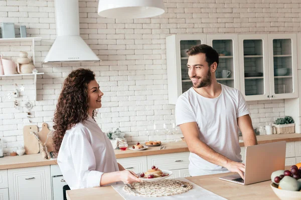 Sonriente pareja con portátil y sabrosos panqueques mirándose en la cocina - foto de stock