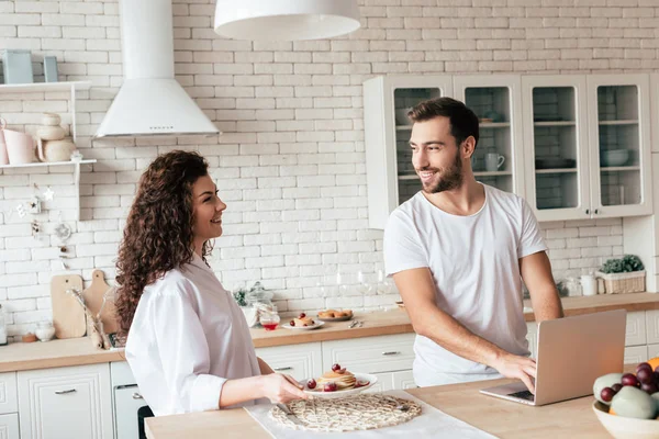 Coppia sorridente con computer portatile e gustosi pancake guardarsi in cucina — Foto stock