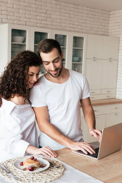 Sonriente pareja usando portátil durante el desayuno en la cocina - foto de stock