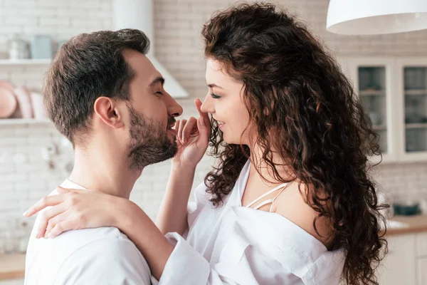 Улыбающаяся любящая пара в белой одежде, обнимающая кухню — стоковое фото