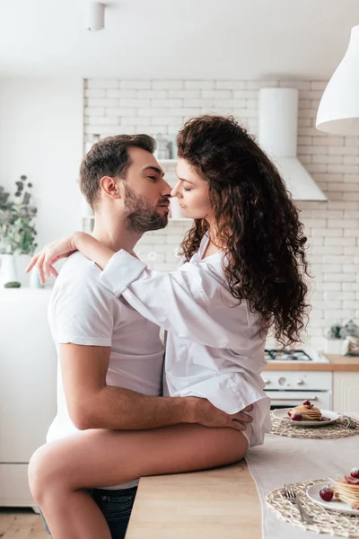 Sexy girl sitting on table and embracing with boyfriend in kitchen — Stock Photo