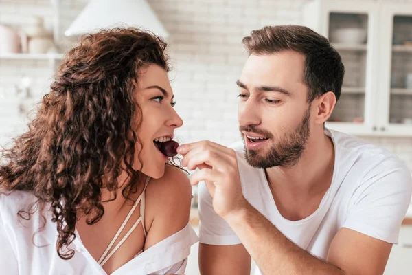 Bearded handsome man feeding girlfriend with berry in kitchen — Stock Photo