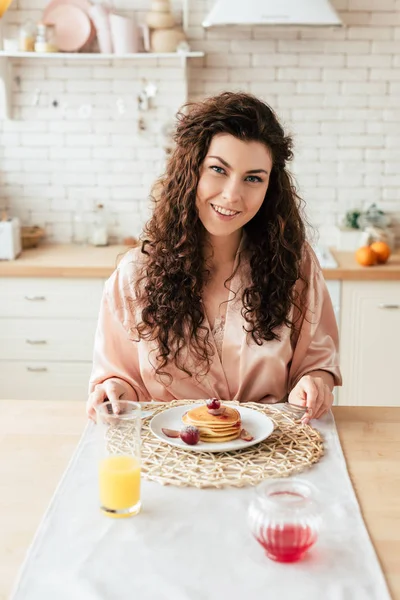 Smiling curly young woman sitting at table with plate of pancakes — Stock Photo