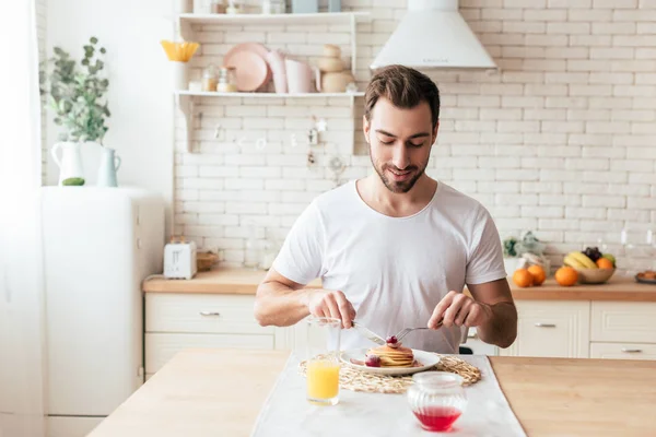 Hombre barbudo sonriente en camiseta blanca comiendo panqueques en la cocina - foto de stock
