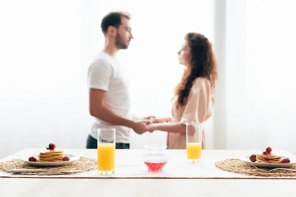 Foco seletivo do casal de mãos dadas com panquecas e suco de laranja em primeiro plano — Fotografia de Stock