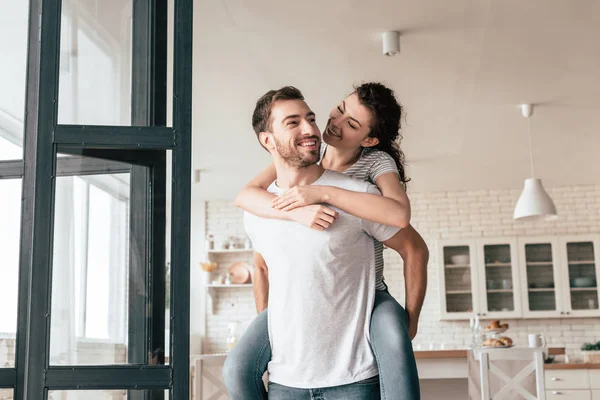 Rindo barbudo homem carregando namorada piggyback em casa — Fotografia de Stock