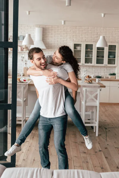 Ridere uomo barbuto portando fidanzata a cavalluccio in cucina — Foto stock