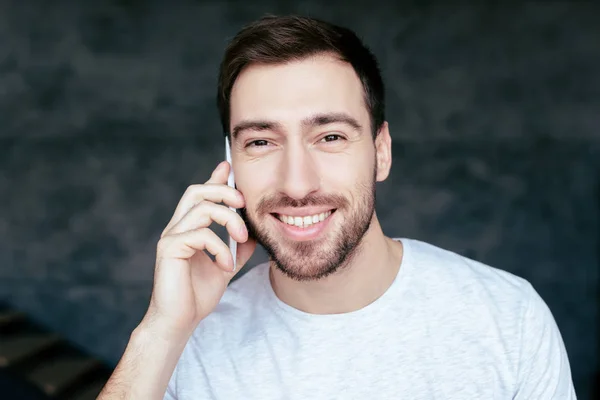 Vista frontal del hombre barbudo sonriente hablando en el teléfono inteligente y mirando a la cámara - foto de stock