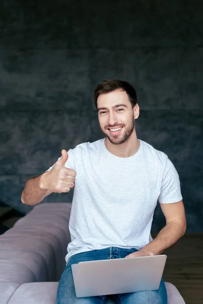 Laughing man using laptop and showing thumb up in living room — Stock Photo