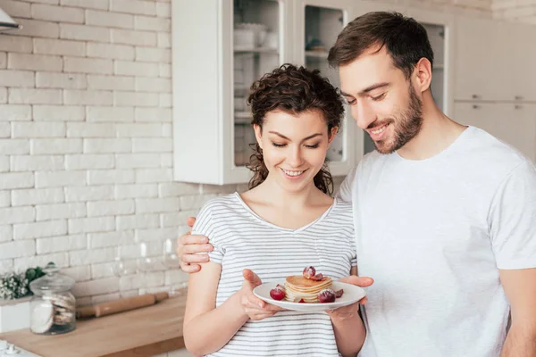 Sonriente hombre abrazando novia y mirando panqueques en la cocina - foto de stock