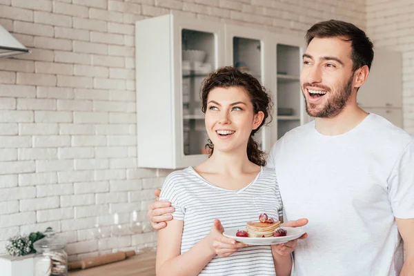 Sonriente hombre abrazando novia y mirando hacia otro lado en la cocina - foto de stock