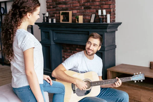 Homme barbu souriant jouant de la guitare acoustique à petite amie dans le salon — Photo de stock