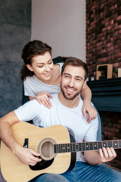 Улыбающаяся девушка обнимает парня, пока он играет на акустической гитаре — стоковое фото
