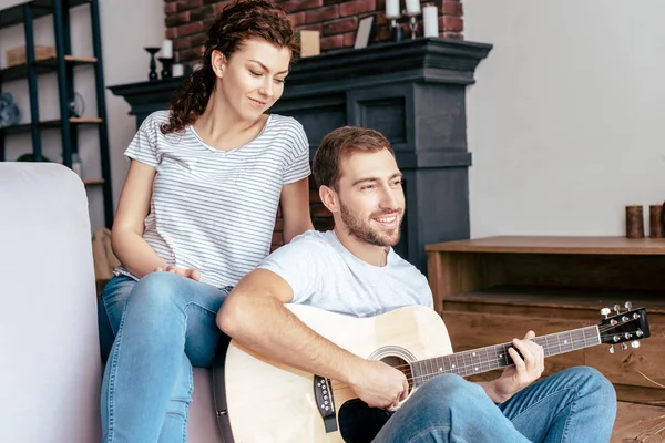 Uomo barbuto sorridente suonare la chitarra acustica alla fidanzata in soggiorno — Foto stock