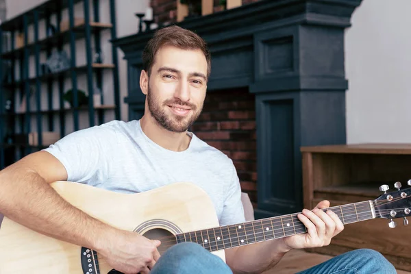Uomo barbuto sorridente che suona la chitarra acustica in salotto — Foto stock
