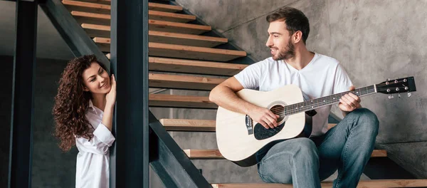 Plan panoramique d'un homme souriant assis sur les escaliers et jouant de la guitare acoustique à sa petite amie — Photo de stock