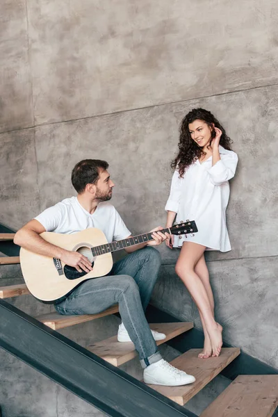 Sonriente hombre barbudo sentado en las escaleras y tocando la guitarra acústica a su novia - foto de stock