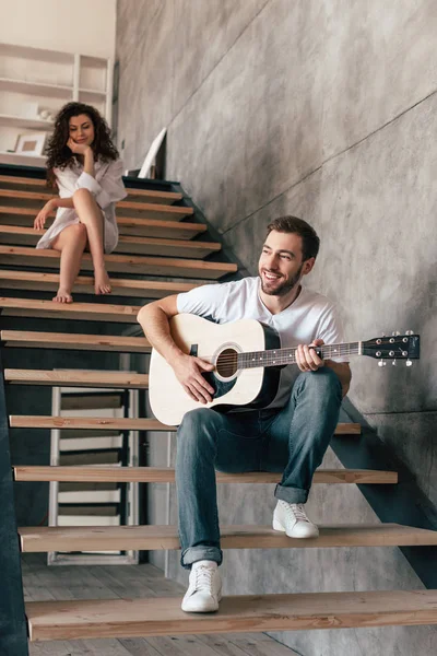 Улыбающийся бородатый мужчина, сидящий на лестнице и играющий на акустической гитаре с девушкой — стоковое фото