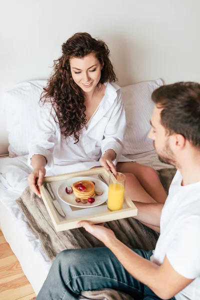 Sonriente pareja sentada en la cama y sosteniendo bandeja con desayuno - foto de stock