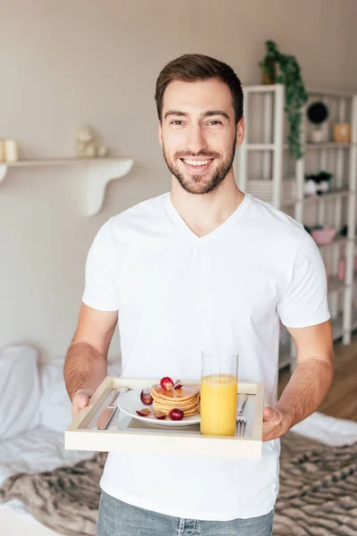 Smiling man holding tray with pancakes and glass of orange juice in bedroom — Stock Photo