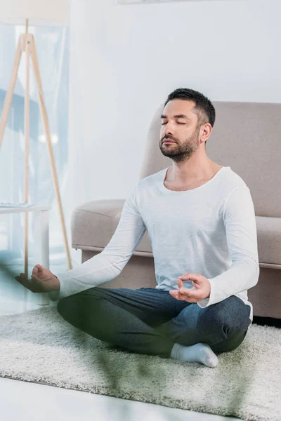 Селективное внимание красивого мужчины, сидящего на ковре в Lotus Pose и медитирующего дома — стоковое фото