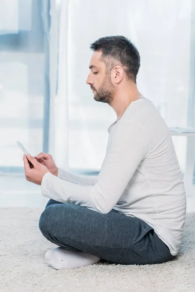 Vista lateral del hombre guapo en ropa casual sentado en la alfombra y el uso de teléfono inteligente en casa - foto de stock