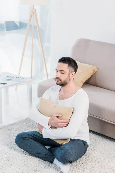 Hombre guapo con los ojos cerrados sentado en la alfombra y abrazando almohada en casa en la sala de estar - foto de stock