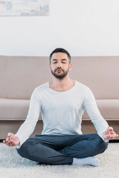 Hombre guapo con los ojos cerrados sentado en la alfombra en Lotus Pose y meditando en casa - foto de stock