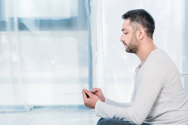 Side view of handsome man in casual clothes sitting on carpet and using smartphone at home with copy space — Stock Photo