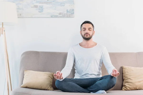 Handsome man with eyes closed sitting on couch in Lotus Pose and meditating at home — Stock Photo