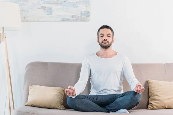 Красивый мужчина с закрытыми глазами сидит на диване в Lotus Pose и медитирует дома — стоковое фото