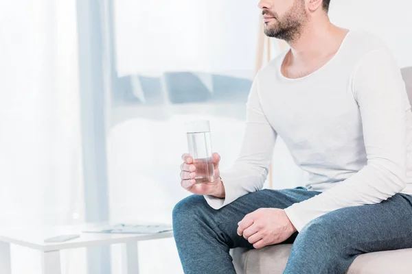 Cropped view of bearded man sitting on couch and holding glass of water at home with copy space — Stock Photo