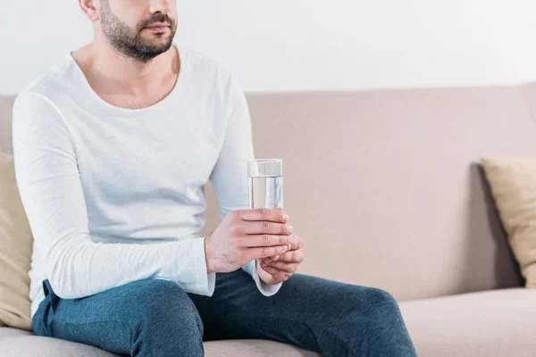 Cropped view of bearded man sitting on couch and holding glass of water at home with copy space — Stock Photo