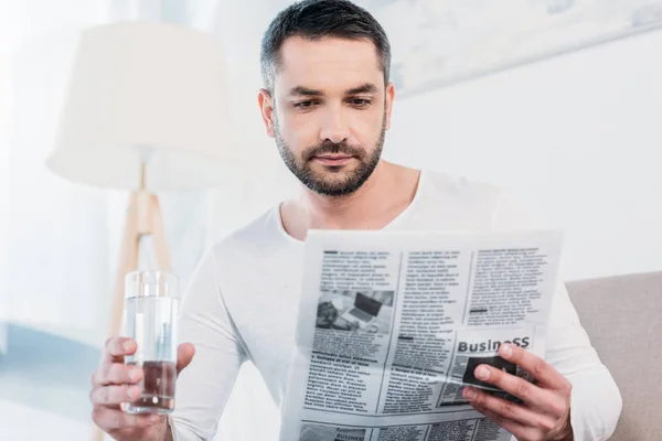 Bell'uomo barbuto seduto con un bicchiere d'acqua e leggendo giornale a casa — Foto stock