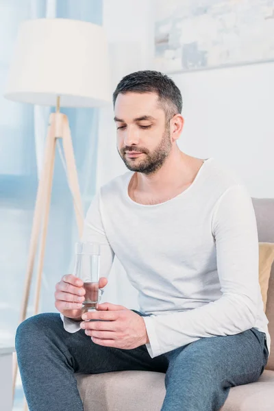 Hombre barbudo guapo sentado en el sofá y sosteniendo un vaso de agua en casa - foto de stock