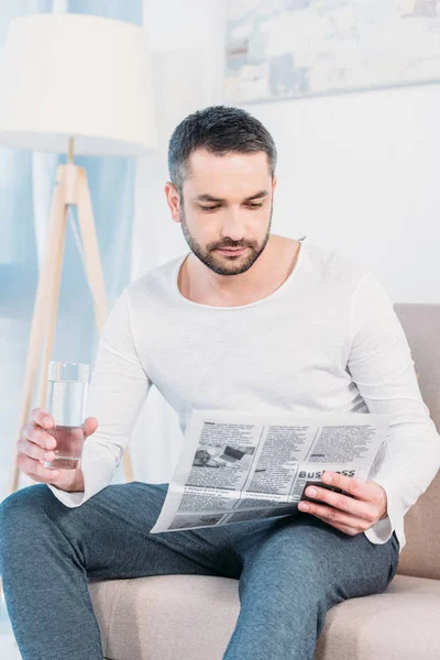 Bonito barbudo homem sentado no sofá com vidro de água e ler jornal em casa — Fotografia de Stock