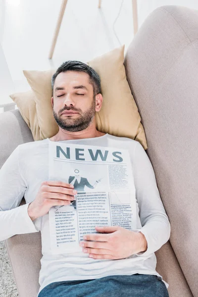 Hombre barbudo guapo con los ojos cerrados sosteniendo periódico y descansando en el sofá en casa - foto de stock